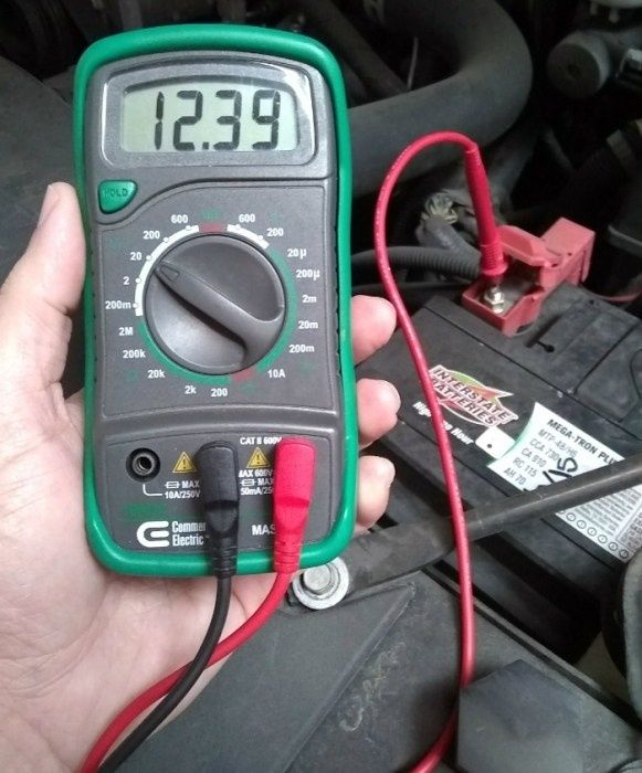 Multimeter zeigt 12,39 Volt an einer Autobatterie im Motorraum